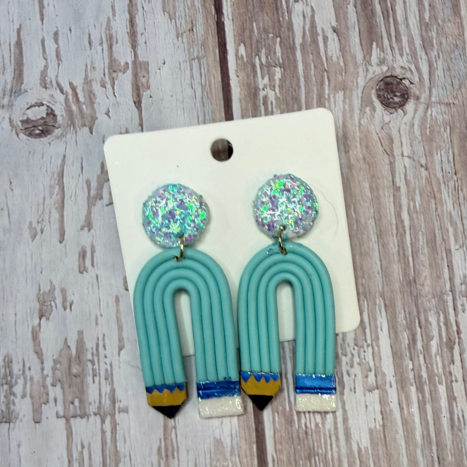 A pair of handmade earrings displayed on a white backing card, placed on a rustic, whitewashed wooden surface. The earrings feature round, glittery silver-blue studs and U-shaped turquoise pencils with whiteand blue striped pencil tips.