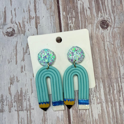 A pair of handmade earrings displayed on a white backing card, placed on a rustic, whitewashed wooden surface. The earrings feature round, glittery silver-blue studs and U-shaped turquoise pencils with whiteand blue striped pencil tips.