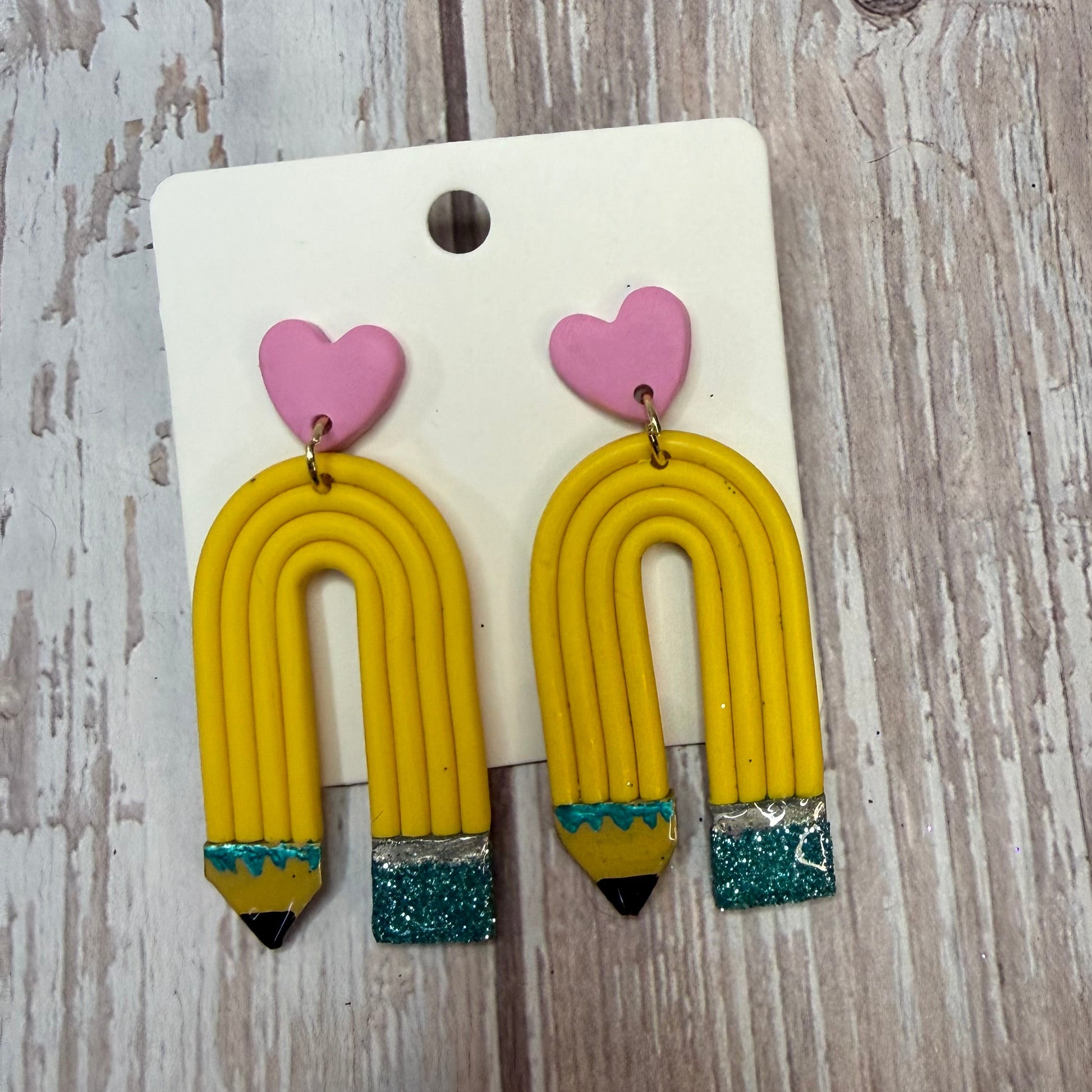 A pair of yellow pencil-shaped earrings with pink heart charms and glittery teal eraser  tips, displayed on a white backing card over a rustic wooden surface.